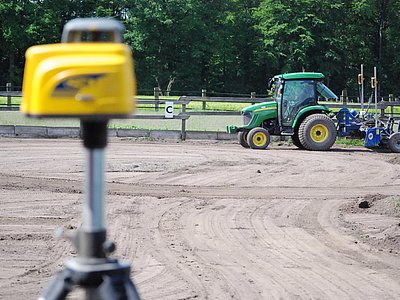 Vorbereitung der Tragschicht im Reitplatzbau mit Lasernivellierung im Vordergrund und Traktor mit Planiergerät im Hintergrund